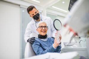 Older lady patient admiring dental implants in mirror sitting in dentist's chair.
